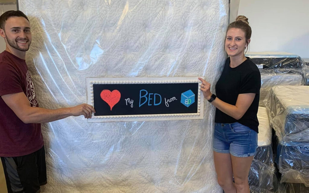 Happy customers holding a sign that says 'I love my bed from BoxDrop Billings' during mattress shopping in Billings, MT.